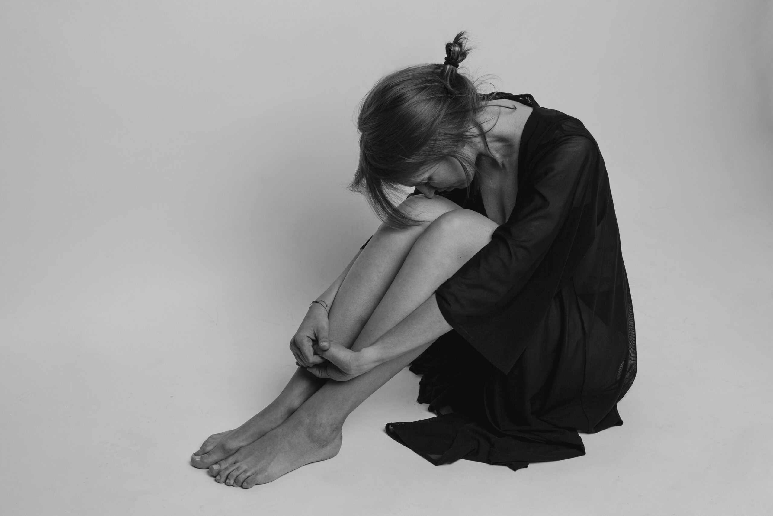A black and white image of a young woman in a black dress sitting mournfully, curled into a ball against a light gray background
